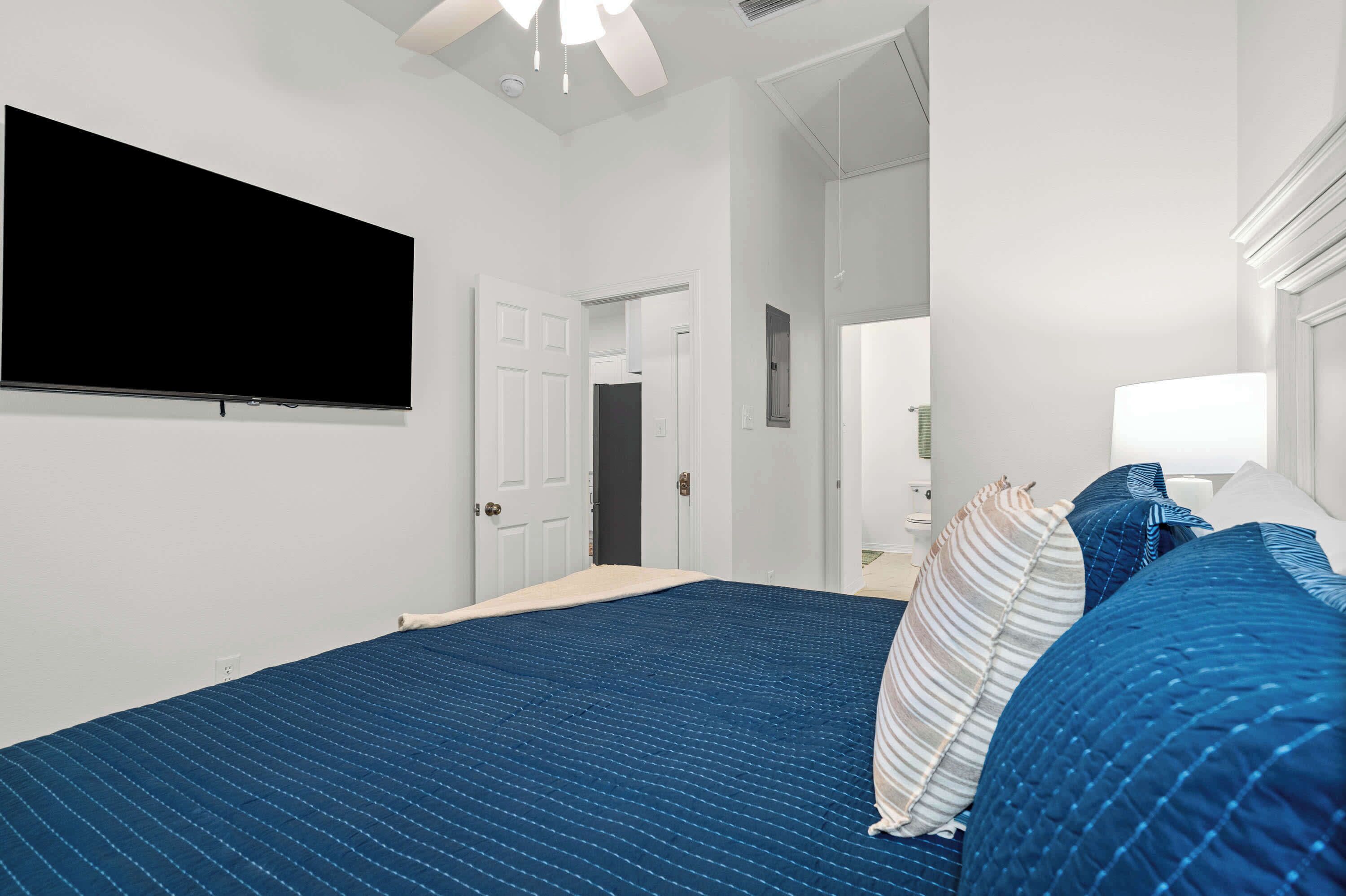 Une chambre dotée d’un grand lit, d’un téléviseur à écran plat fixé au mur, d’une porte donnant sur une autre pièce et d’un ventilateur de plafond.