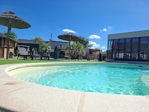 A swimming pool with lounge chairs and umbrellas, surrounded by a fenced yard with palm trees and a house. - Idyllic cottage (Gennes-Val-de-Loire)
