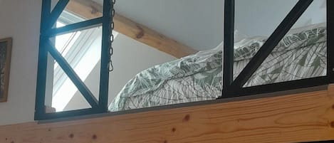 Een loft-stijl kamer met een plafond van houten balken, een hoogslaper met een geometrisch hoofdeinde, een klein fornuis, een trapje en een raam.