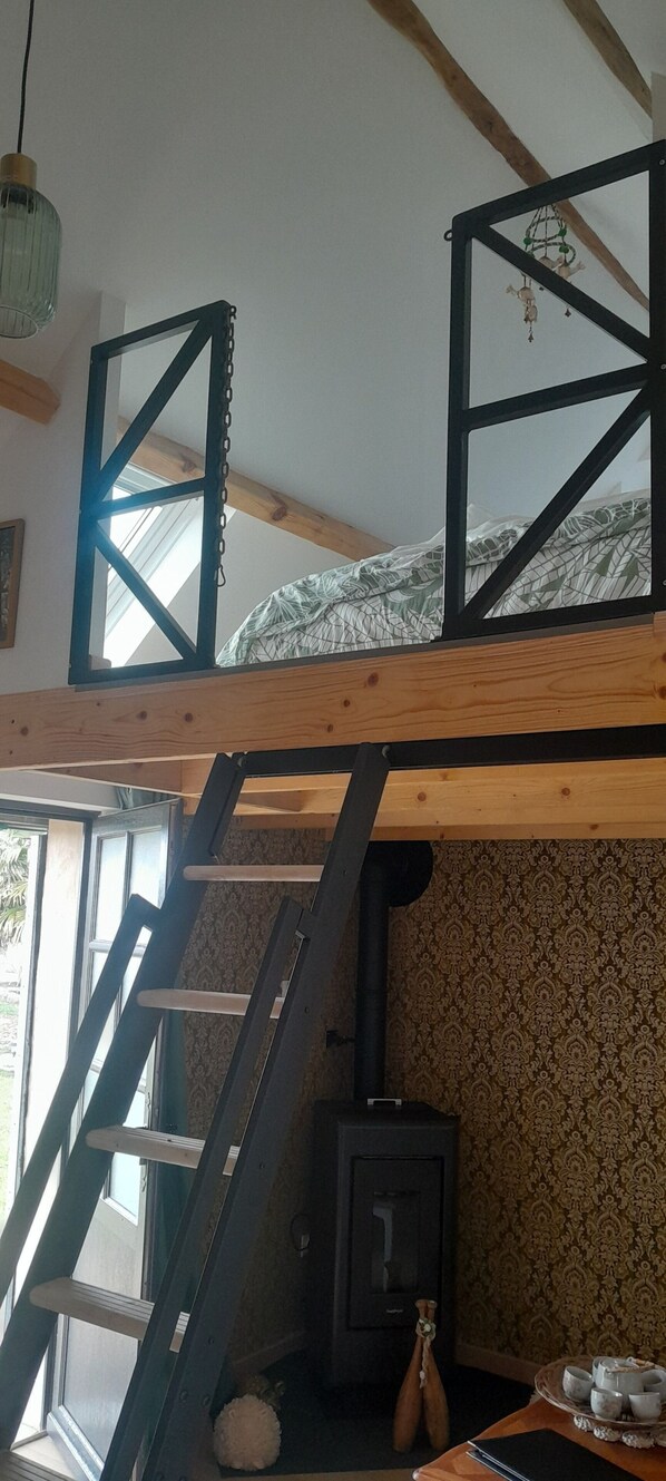Habitación tipo loft con techo de vigas de madera, cama superior con cabecera geométrica, cocina pequeña, escalera y ventana.