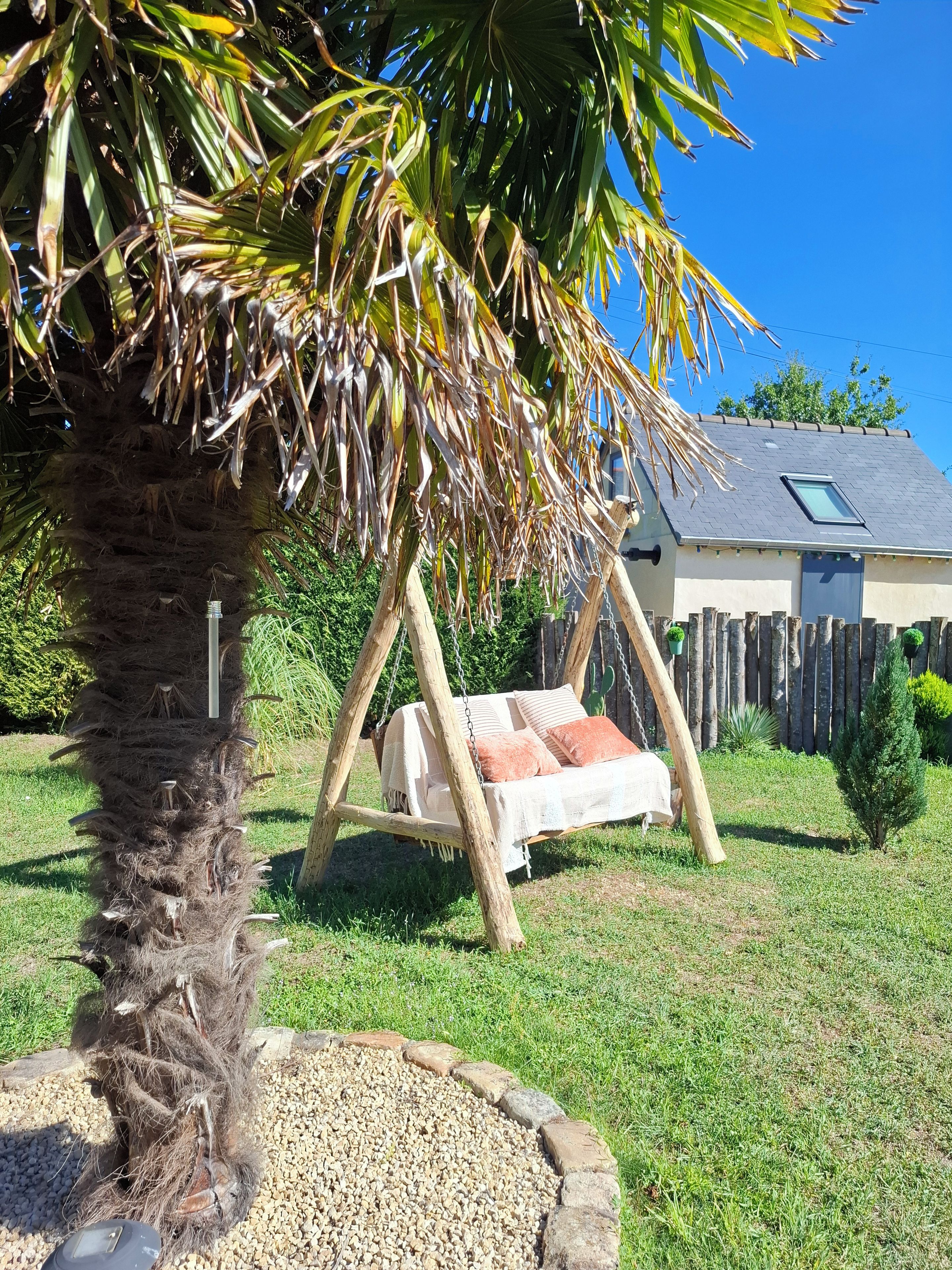 A garden with a swing chair, a palm tree, and a house in the background.