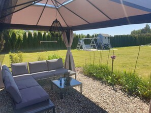 An outdoor seating area with a sofa and table, a pergola overhead, and a playground in the background. - Villa Near Baltic Sea (2.5 km) – 4 Bedrooms, 3 Bathrooms, Private Garden (Łebcz)