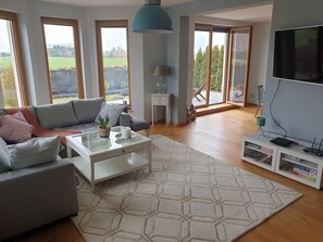 A modern living room with a sofa, coffee table, and TV. Large windows offer views of greenery and a distant landscape. - Villa Near Baltic Sea (2.5 km) – 4 Bedrooms, 3 Bathrooms, Private Garden (Łebcz)