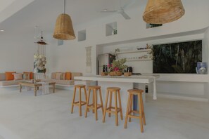 A modern living room with a white sofa, wooden stools, a white coffee table, wicker pendant lights, a large TV, and a wall-mounted shelf with decorative items.
