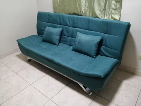 A teal sofa with two cushions in a room with white tiled flooring.. Shop-Changuinola-Fit5people