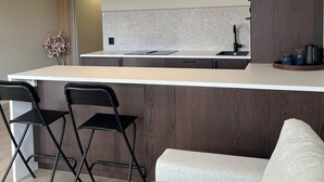 Ett modernt kök med en vit bänkskiva, mörka träskåp och en inbyggd ugn. I förgrunden står två svarta stolar och en beige soffa.