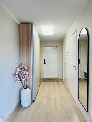 A hallway with a wooden floor, a white vase with pink flowers, a large mirror, and a door at the end. - Apartment in Moss city center (Østfold)