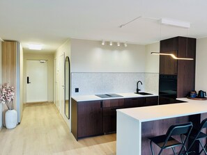 A modern kitchen with a central island, a sink, and a stove. There are wooden cabinets and a white countertop. A vase with flowers is on the left, and a chair is at the island. - Apartment in Moss city center (Østfold)