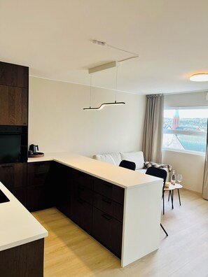 A modern kitchen with a central island, dark wooden cabinets, and a view of a cityscape through the window. - Apartment in Moss city center (Østfold)
