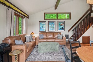 Un salon avec deux canapés en cuir marron, un pouf vert, un tapis à motifs et une table à manger en bois avec des chaises.