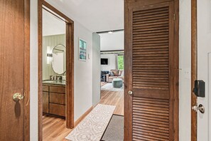 Un pasillo con piso de madera, una alfombra estampada, una puerta con mirilla y vista a una sala de estar con sofá y televisor.