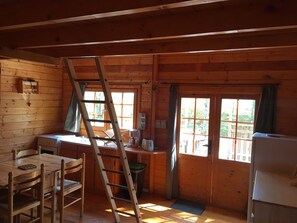 LâintĂ©rieur dâune cabane en bois comprend une table Ă manger, des chaises, une Ă©chelle, une petite kitchenette et une fenĂȘtre avec des rideaux.
