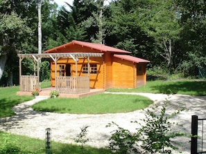 A wooden cabin with a porch, surrounded by a gravel path and greenery.