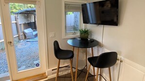 A small room with a door leading to an outdoor area, a round table with two chairs, a television mounted on the wall, and a potted plant on the table.