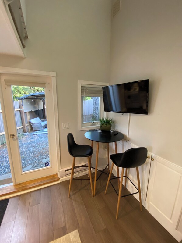 A small room with a door leading to an outdoor area, a round table with two chairs, a television mounted on the wall, and a potted plant on the table.