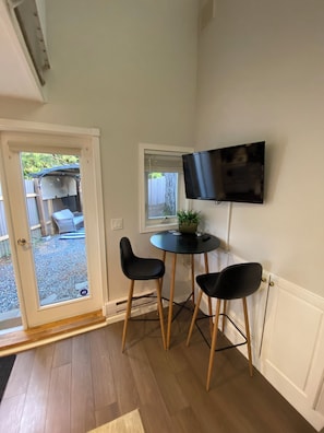 A small room with a door leading to an outdoor area, a round table with two chairs, a television mounted on the wall, and a potted plant on the table.
