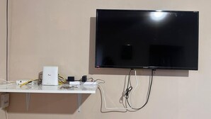 En platt-TV som är monterad på en vägg, med en vit hylla under som innehåller elektroniska apparater och kablar.