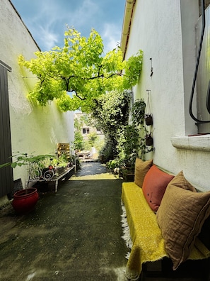 Une petite cour intérieure avec un canapé jaune, des plantes en pot et un arbre qui offre de l’ombre.