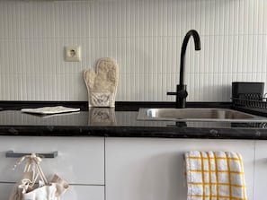 A kitchen with a black faucet, a white tiled backsplash, a stainless steel sink, a white oven mitt, and a towel rack with a towel. - * Coastal Paradise: Your relaxing corner by the beach (O Pindo)