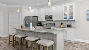 Una cocina moderna con armarios blancos, una isla de granito con taburetes altos, electrodomésticos de acero inoxidable y un salpicadero con azulejos en patrón de espiga.