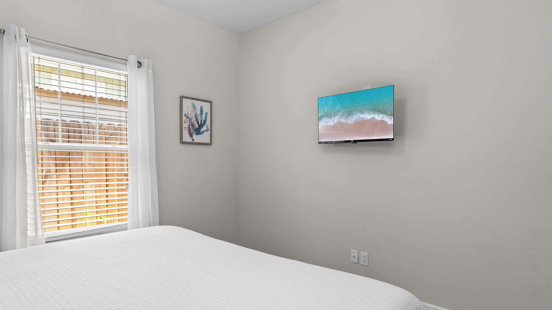 A bedroom with a bed, a window with blinds, a wall-mounted TV, and a framed picture.