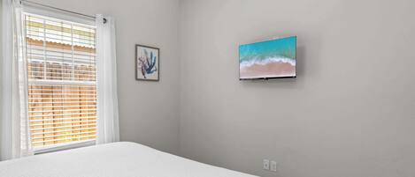 A bedroom with a bed, a window with blinds, a wall-mounted TV, and a framed picture.