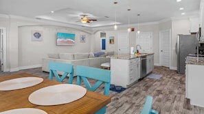 Eine moderne Küche mit Kochinsel, ein Essbereich mit Holztisch und blauen Stühlen sowie ein Wohnzimmer mit grauem Sofa.
