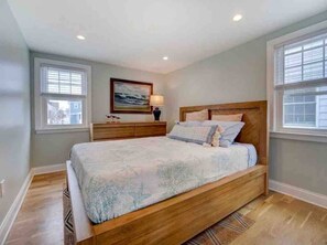 Un dormitorio con cama, mesitas de noche, un cuadro y ventanas.
