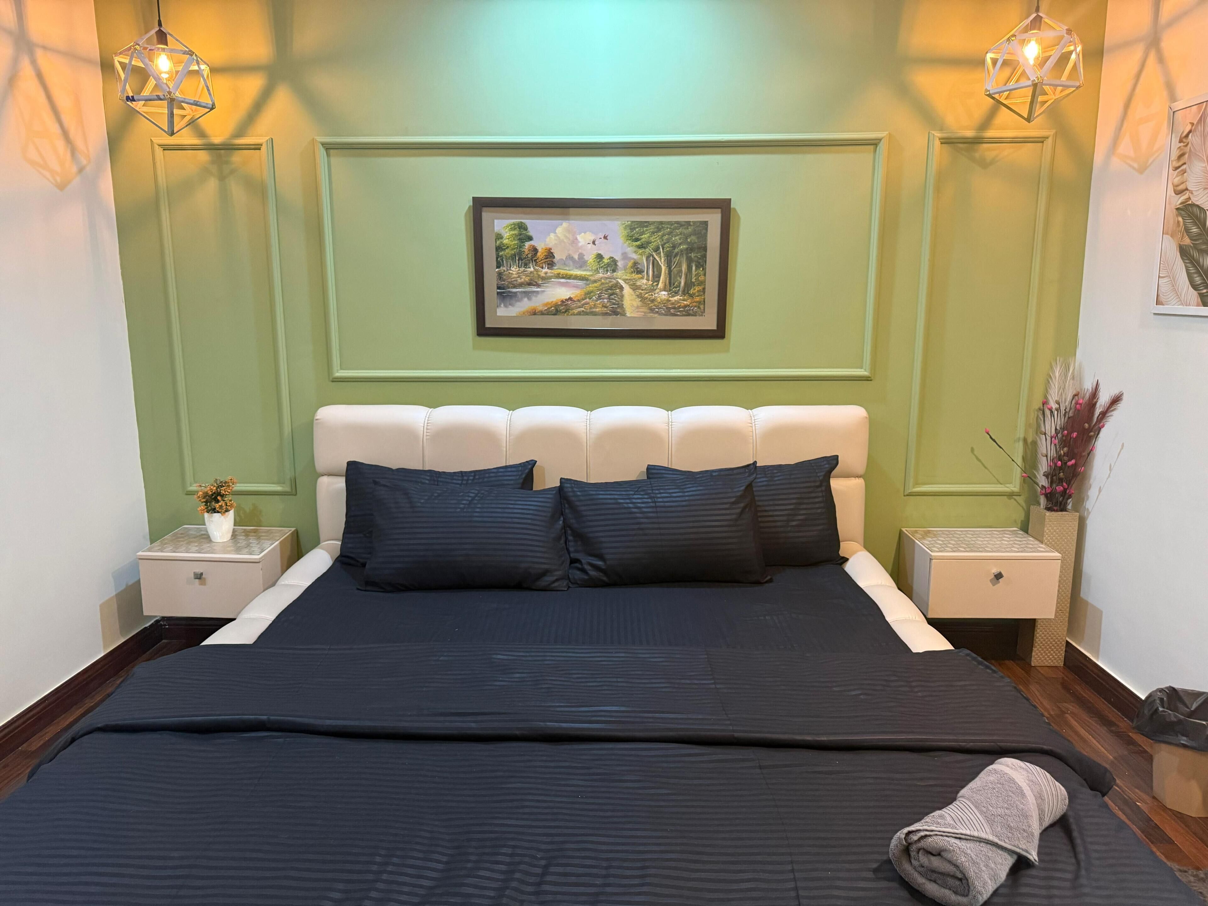 Een netjes opgemaakt bed met een donkere sprei, twee kussens en een opgevouwen handdoek ernaast. Daarboven hangt een lijst met een landschapsafbeelding en aan weerszijden van het bed zijn twee wandlampen bevestigd.