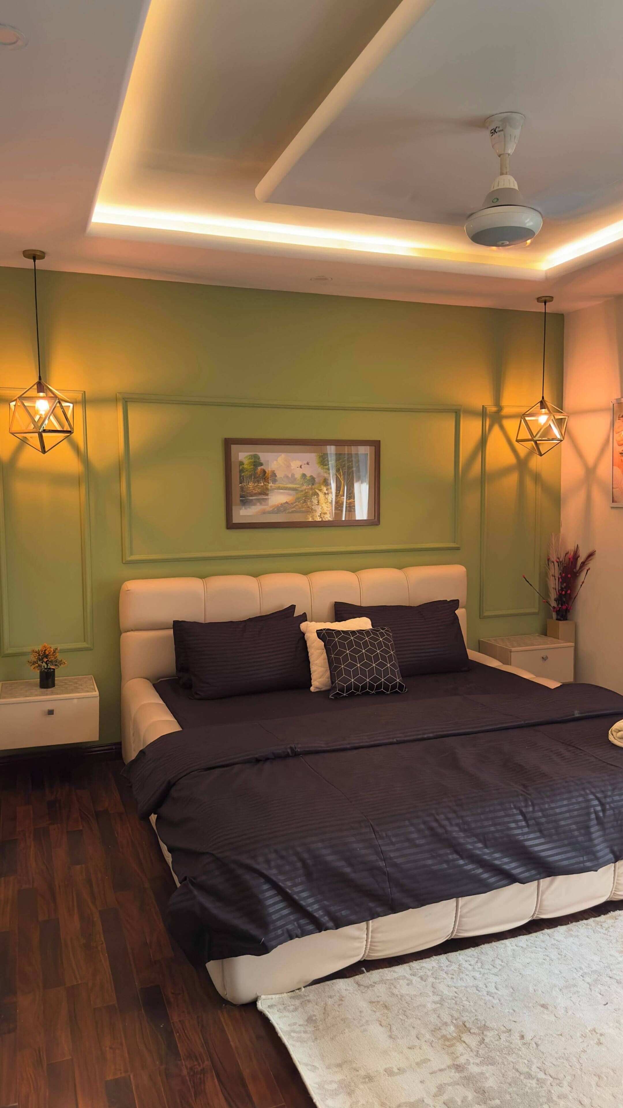 Un dormitorio con cama, mesitas de noche, ventilador de techo e imagen colgada en la pared.