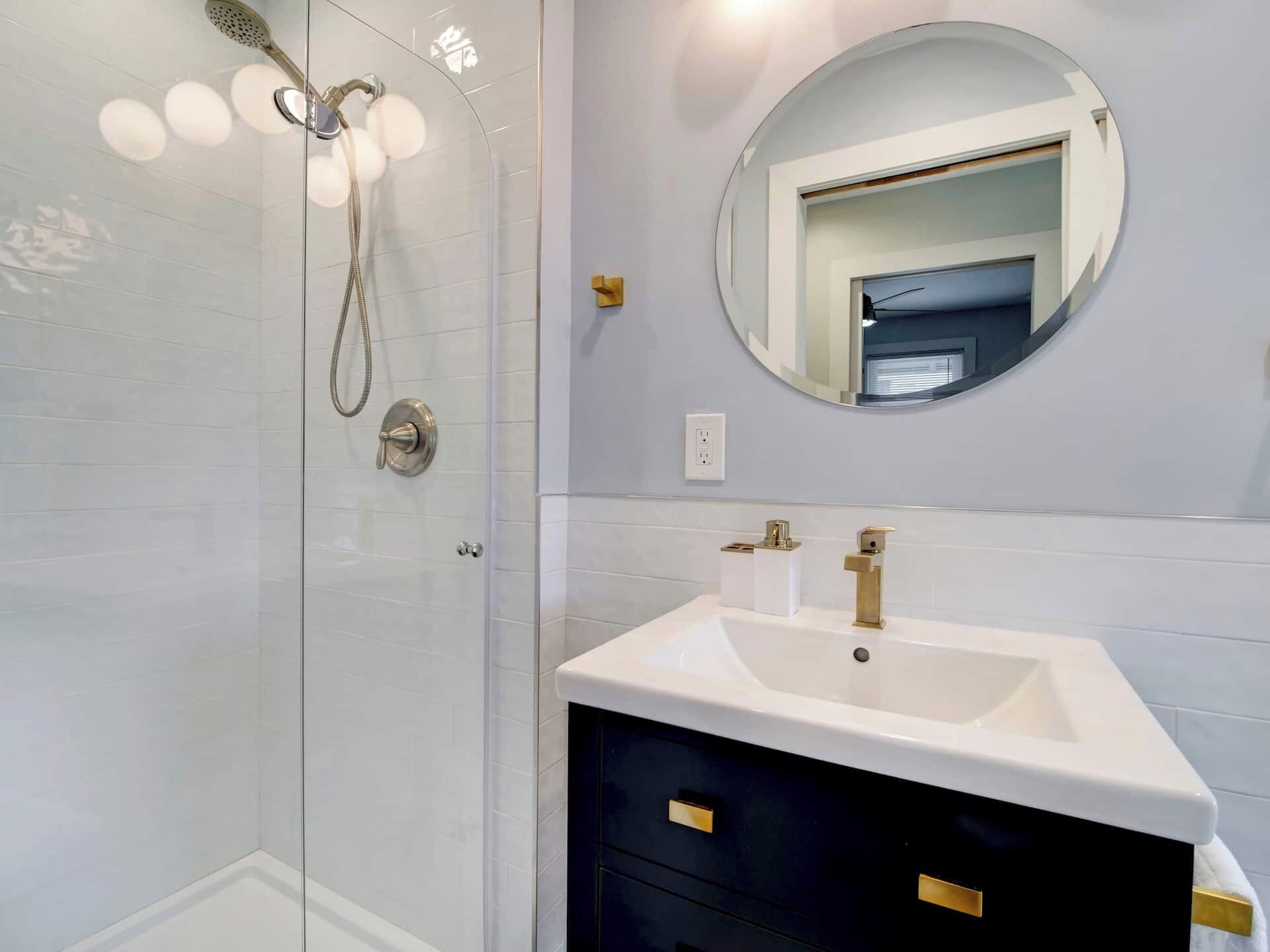 Un baño moderno con cabina de ducha de vidrio, un lavabo blanco con detalles dorados y un espejo redondo en la pared.