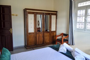 Une chambre d’hôtel comprenant un lit, une armoire en bois, une chaise et une fenêtre ornée de barreaux en fer.