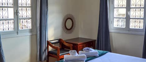 Un dormitorio con cama, escritorio, silla, candelabro y ventanas con cortinas.