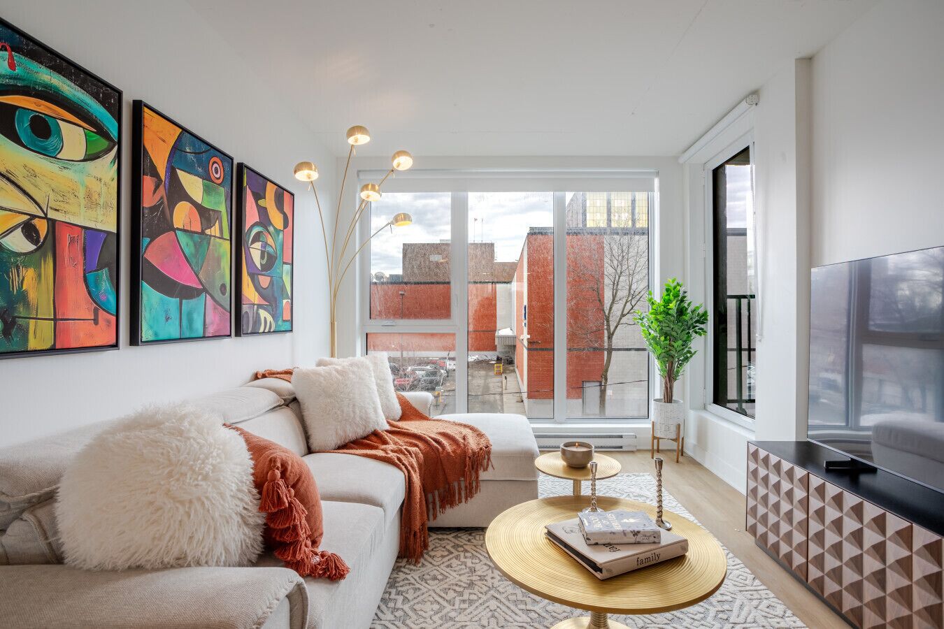 Un salon moderne avec un canapé, une table basse et un téléviseur. Les murs sont ornés de tableaux abstraits et un balcon offre une vue sur les bâtiments.