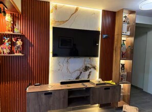 Un salon moderne doté d’un grand téléviseur à écran plat fixé au mur, lequel présente un motif évoquant le marbre, de panneaux en bois sur les côtés et un meuble de rangement intégré comprenant un support pour téléviseur et des étagères.