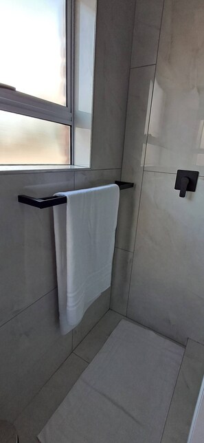 Une salle de bain avec une serviette suspendue à un porte-serviettes noir, un tapis de bain blanc sur le sol et une fenêtre laissant entrer la lumière naturelle.
