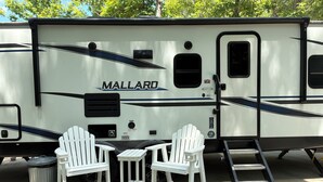 A travel trailer with a 'MALLARD' branding, two white Adirondack chairs, a small table, a trash can, and a ladder. - Gated Luxury Glamping at Diamond Acres – Near Grove Marina & Cumberland Falls (Corbin)