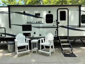 A travel trailer with a 'MALLARD' branding, two white Adirondack chairs, a small table, a trash can, and a ladder.