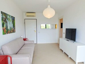 Una sala de estar con un sofá, un televisor sobre un mueble blanco, un cuadro en la pared y una puerta con ventana.