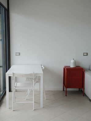 Una habitación minimalista con una mesa y sillas blancas, una mesita auxiliar roja con una lámpara, y un sofá gris.