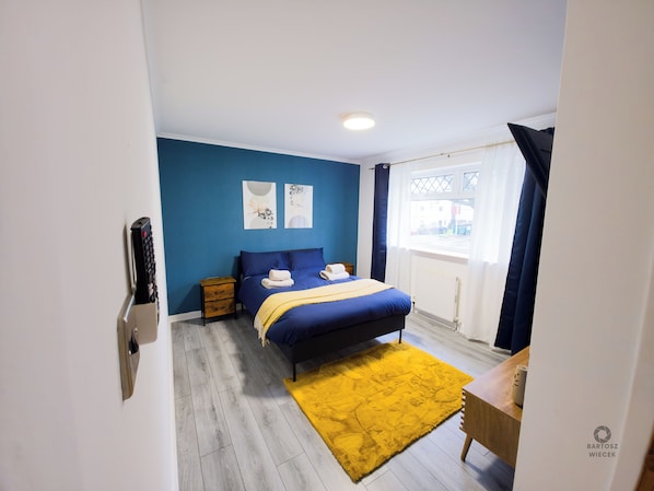 Une chambre à coucher avec un lit, une table de chevet en bois, un tapis jaune et une fenêtre avec des rideaux.