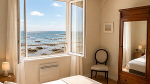 A bedroom with a bed, a chair, a wooden wardrobe, a window with a view of the sea, and a bedside lamp. - LocaLise - La clé de l'Océan - House with exceptional sea views (Treffiagat)