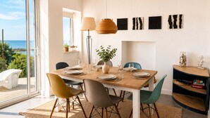 A dining area with a wooden table set for four, surrounded by chairs, a vase with a plant, and a wall with abstract art. - LocaLise - La clé de l'Océan - House with exceptional sea views (Treffiagat)