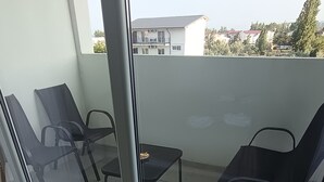 A balcony with two chairs and a small table, a glass door, and a view of buildings and trees. - Roma Apartment luxury flat in a beautiful location in Mamaia Beach by the Sea. (Năvodari)