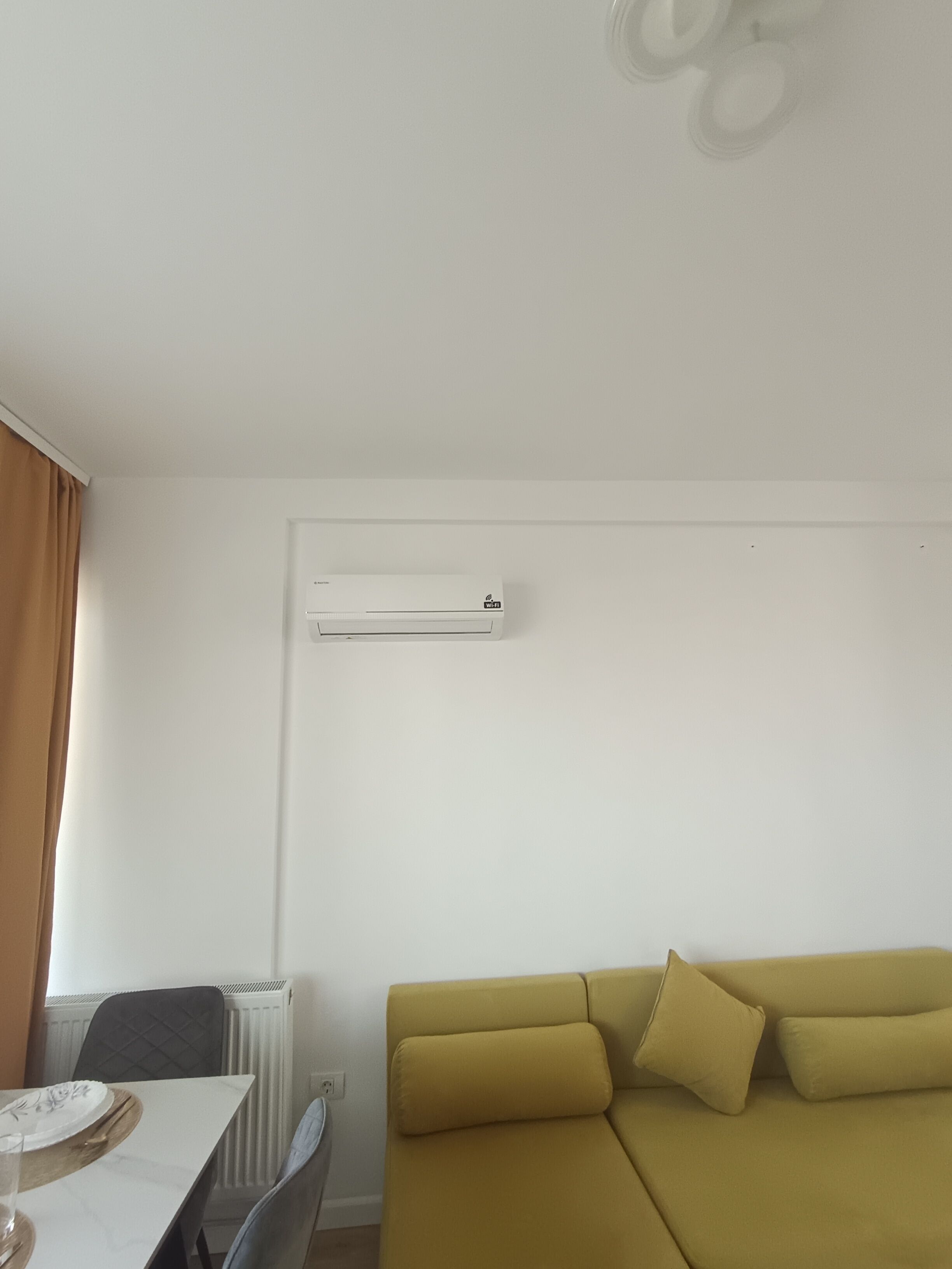 A room with a white air conditioner, a yellow sofa, a dining table with a chair, and a radiator.