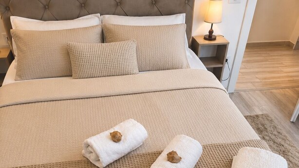 A neatly made bed with a tufted headboard, a beige quilt, and three rolled towels with brown decorative ends. - Roma Apartment luxury flat in a beautiful location in Mamaia Beach by the Sea. (Năvodari)