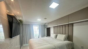 A modern bedroom with a large bed, a TV mounted on the wall, and a window with curtains. - Blue Sunset Cabo Branco | Beachfront Apartment (João Pessoa)