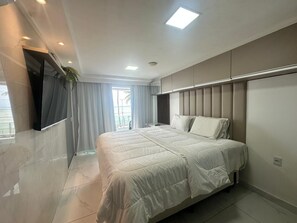 A modern bedroom with a large bed, a TV mounted on the wall, and a window with curtains. - Blue Sunset Cabo Branco | Beachfront Apartment (João Pessoa)