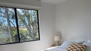 A bedroom with a large bed, a window with a view of trees, a bedside lamp, and a wooden floor. - Beach Stay | Walk to the Surf, Café & Shops (Coolum Beach)