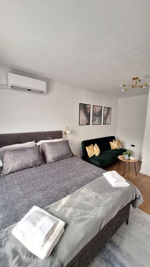 A modern bedroom with a large bed, a green sofa, a small side table, and a wall-mounted air conditioner.